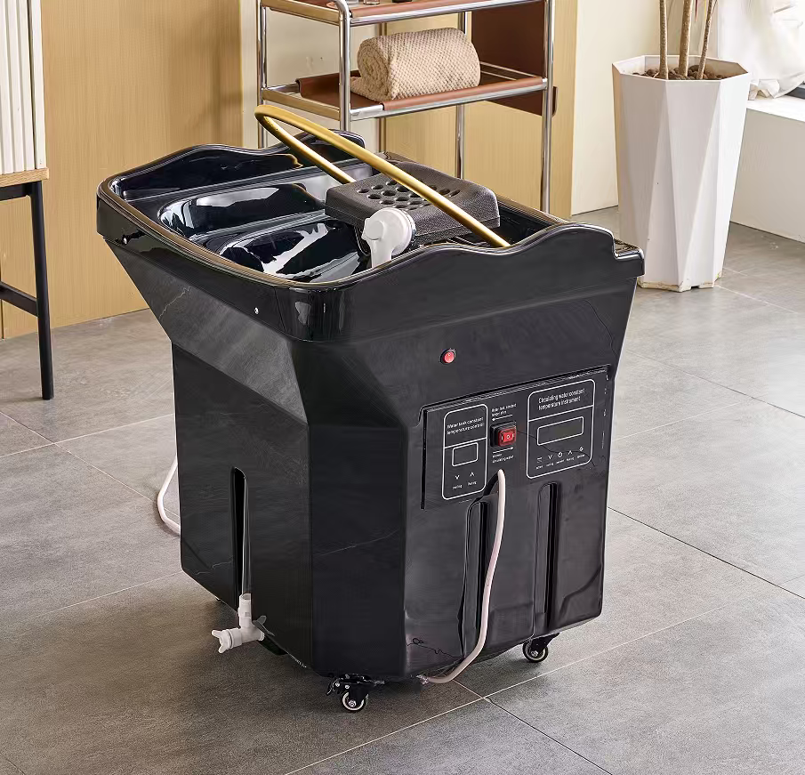 Black portable pedicure spa basin on wheels with control panels on the side in a salon setting, hose connections visible.